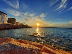 the sun is setting over the water near some rocks and buildings on the shore line