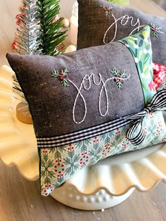 two decorative pillows on a plate with christmas tree decorations in the back and joy written on them