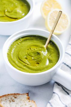 two white bowls filled with green soup next to slices of bread and lemon wedges