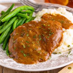 two pieces of meat covered in gravy on a plate with mashed potatoes and green beans