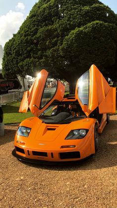 an orange sports car parked on top of a dirt field next to a tree and grass covered park