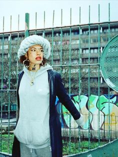 a woman wearing a white hat standing in front of a fence with graffiti on it