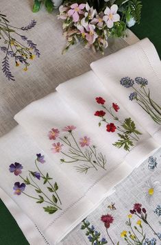 four napkins with flowers on them sitting next to a vase filled with wildflowers