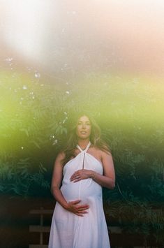 a pregnant woman standing in front of a bush with her hands on her stomach, wearing a white dress