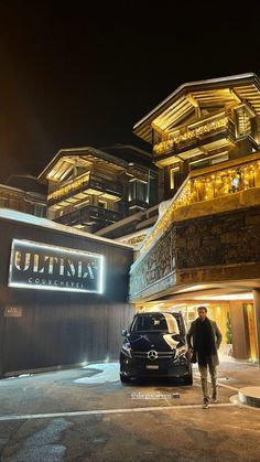 a man walking towards a car in front of a building with lights on it at night