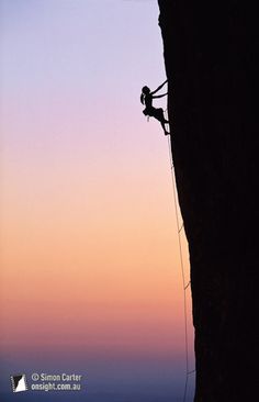 a man climbing up the side of a cliff at sunset with his back to the camera