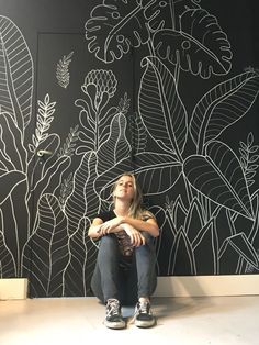 a woman sitting on the floor in front of a wall with plants drawn on it