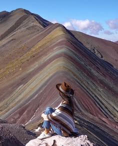 a person sitting on top of a rock next to a rainbow colored mountain range in peru