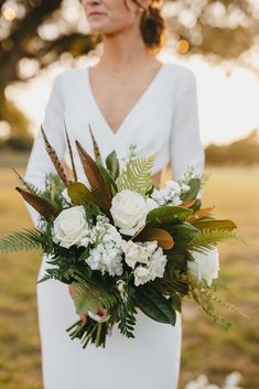 a woman in a white dress holding a bouquet of flowers and greenery on her wedding day