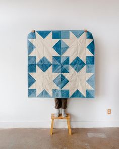 a person standing on a stool holding up a blue and white quilt that is hanging from the wall