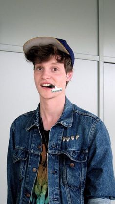 a young man with a toothbrush in his mouth wearing a denim jacket and cap