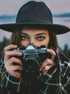 a woman wearing a black hat and holding a camera in front of her face while looking at the camera