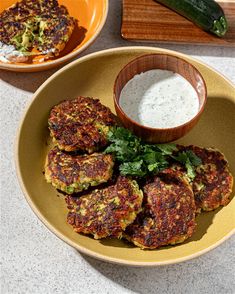 several crab cakes on a plate with sauce and cucumber next to it in a bowl