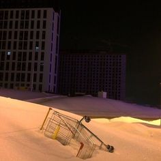 an abandoned shopping cart in the snow at night with buildings in the background and lights on