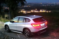 a white bmw suv parked on the side of a road at night with city lights in the background