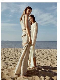 two women standing on top of a sandy beach next to the ocean and one is wearing a white dress