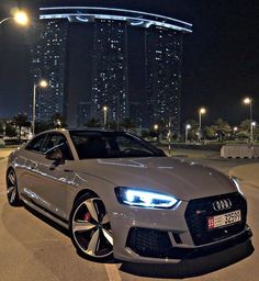 a white car parked in front of a tall building at night with city lights behind it