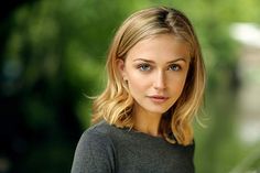 a woman with blonde hair and blue eyes posing for a photo in front of trees