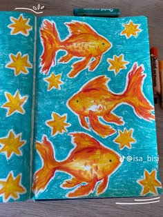 an open notebook with goldfish and stars on it, next to crayon markers