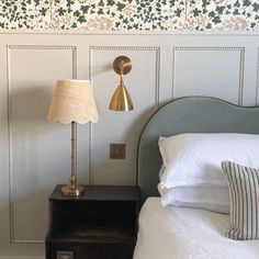 a lamp on a nightstand next to a bed in a room with white sheets and wallpaper