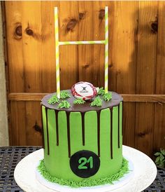 a green cake with chocolate drips and a football on top is sitting on a table in front of a wooden fence