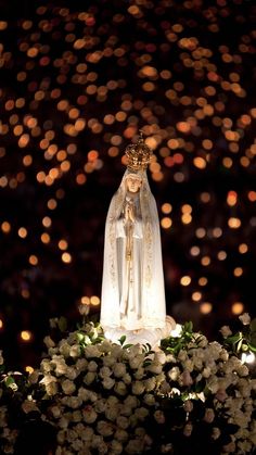 a statue of the virgin mary on top of flowers in front of a lighted background