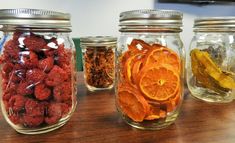 four jars filled with different types of fruits and nuts on a table top, one containing dried oranges, the other unpeeled raspberries