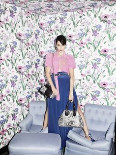 a woman standing in front of a floral wallpaper holding a handbag and purse