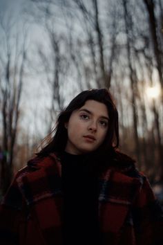 a woman standing in front of trees wearing a red and black plaid coat with her eyes closed