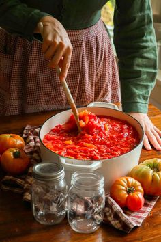 a woman stirring tomato sauce in a pot on a wooden table with tomatoes around it