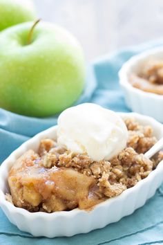 an apple crisp with ice cream in a white bowl on a blue cloth next to two green apples