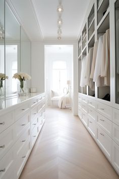 a large white closet with lots of drawers and clothes hanging on the walls, along with vases filled with flowers