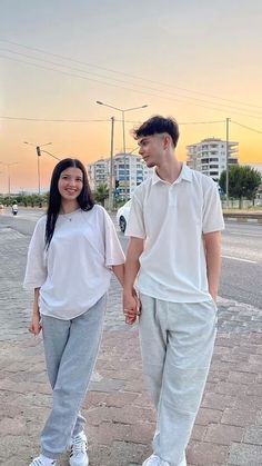 a young man and woman holding hands walking down the street at sunset with buildings in the background