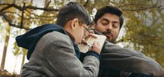 two young men sitting next to each other while talking on cell phones in the park