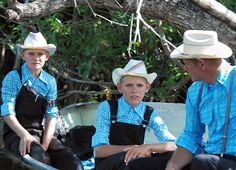 two boys and an older man are sitting on a bench under a tree, wearing cowboy hats