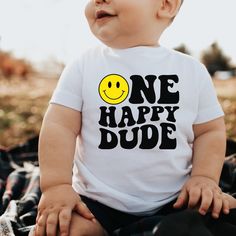 a baby wearing a one happy dude t - shirt while sitting on the ground with his eyes closed