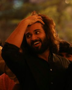 a man with long hair is smiling and holding his hands up to his head while standing in front of other people