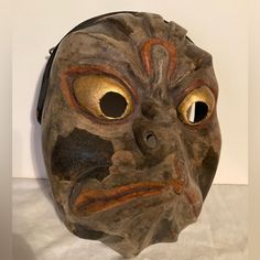 an old mask with big eyes and a face painted on it's side, sitting on a white surface