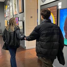 two people holding hands walking through an art gallery with paintings on the wall behind them