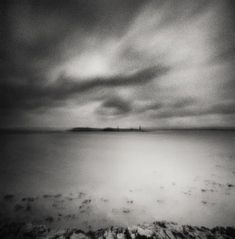 a black and white photo of the ocean with clouds in the sky above it, looking out to sea