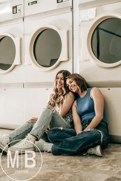 two women sitting next to each other on the ground in front of washers and dryers