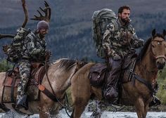 two men riding horses in the water with antlers on their back and one man wearing camouflage