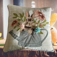 a pillow with an embroidered bunny in a watering can on top of a wooden table