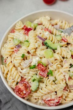 a white bowl filled with pasta salad next to tomatoes and cucumber on the side