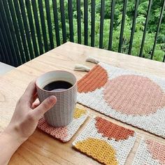 a hand holding a cup of coffee on top of a wooden table next to two crocheted coasters