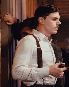 a man with suspenders and a white shirt is looking at his cell phone while standing in front of a door