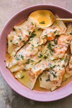 a pink bowl filled with pasta covered in sauce and topped with parsley next to a wooden spoon