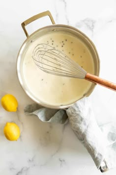 a metal pan filled with liquid next to lemons on a marble counter top and a wooden whisk