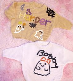 two sweaters that say happy halloween and one with a ghost on the front are laying on a pink blanket