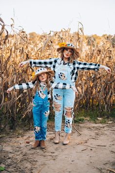 two girls in overalls and cowboy hats standing in front of a cornfield with their arms outstretched